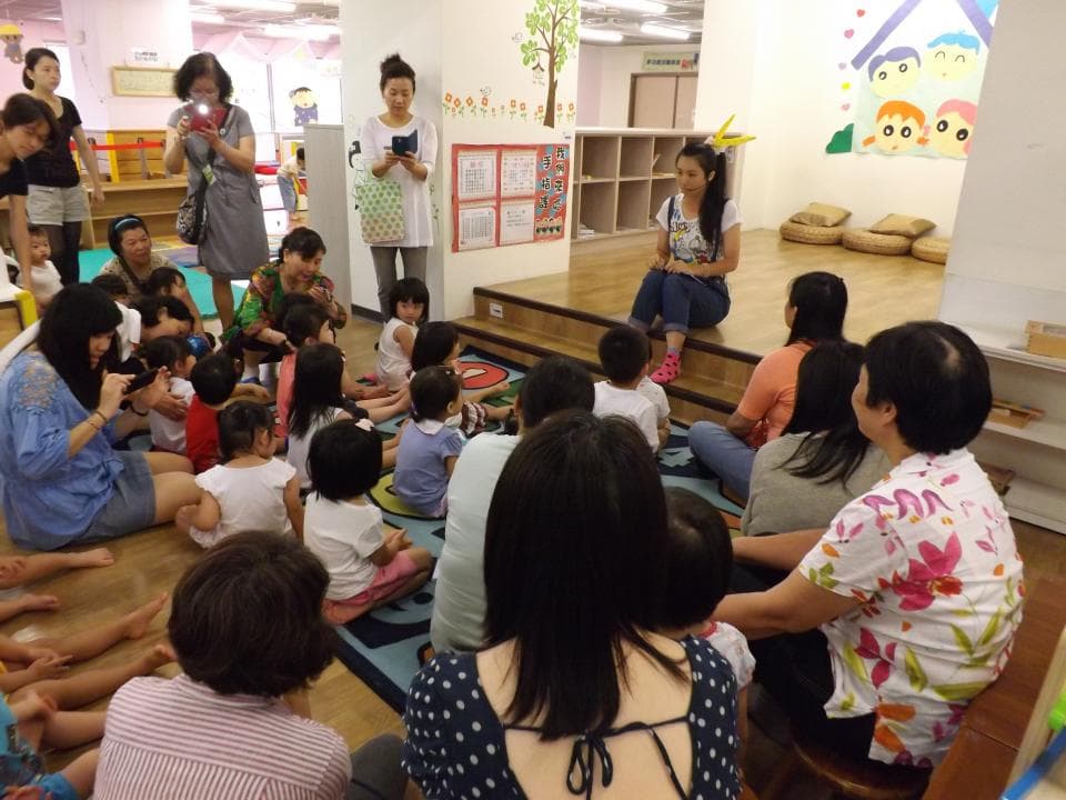 這張照片是一間充滿活力的幼兒園教室,孩子們圍坐在房間中央的地板上,有的在玩耍,有的在聽老師講課。教室裡的牆壁上貼滿了可愛的卡通圖案,充滿了童趣。