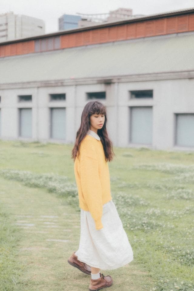 這張「女生在草地上穿著黃色毛衣和白色裙子」的照片,背景是一棟建築物。