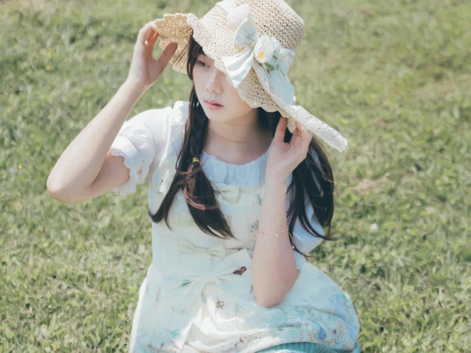 這張照片是一個戴著草帽的少女,她穿著白色蕾絲洋裝,在綠色的草地上。
