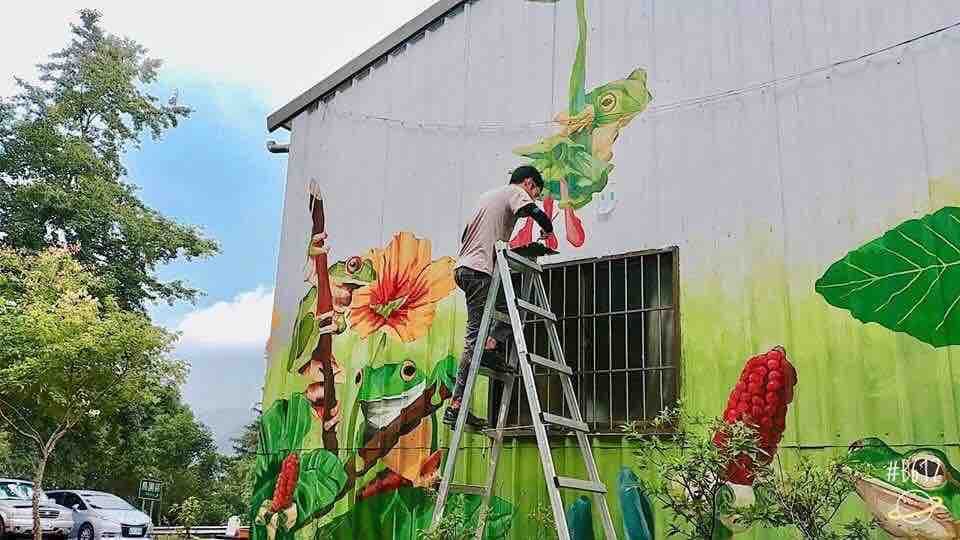 這張照片是一個牆壁上正在創作的壁畫,畫面上有許多青蛙和植物,一名藝術家站在梯子上,仔細地繪製著壁畫。