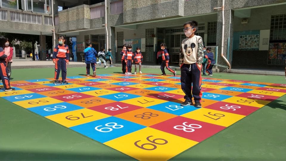 這張照片是一群戴著口罩的小朋友,在戶外玩著一個大型的數字跳格子遊戲。地板上鋪著彩色的方格,每個方格上都寫著不同的數字。小朋友們跳躍在不同的格子之間,看起來非常開心。