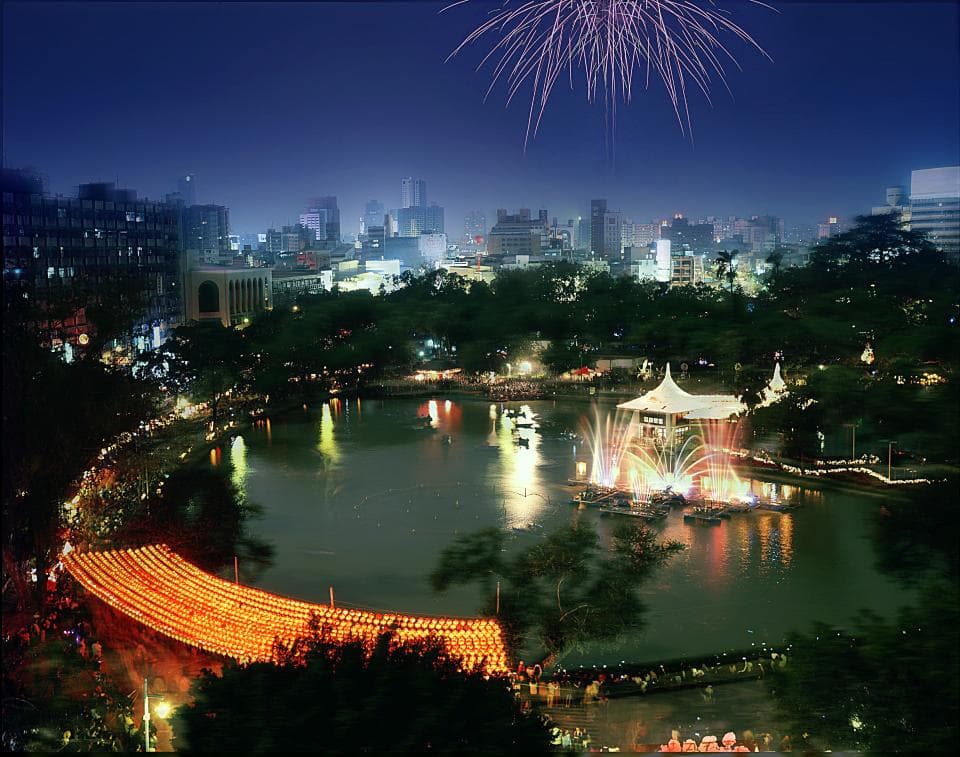 這張「夜晚城市」的照片是,一個城市夜景,夜空有煙火,城市裡有一座湖,湖中有音樂噴泉,湖邊有許多建築。