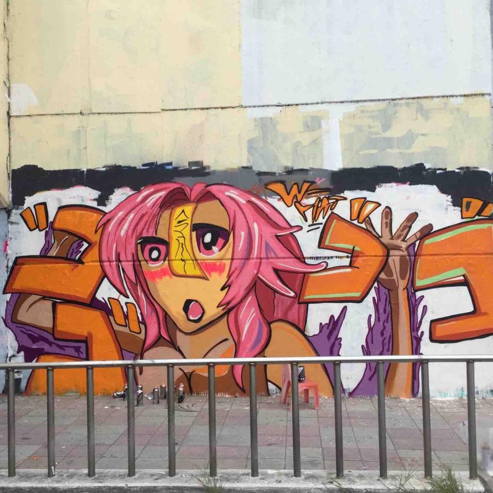 這張照片是塗鴉藝術作品,主要描繪一個紅髮女性角色,她擁有誇張的大眼睛,以及明顯的粉紅色嘴唇。角色被塗鴉在牆上,並以鮮豔的顏色呈現,營造出充滿活力和叛逆的氛圍。