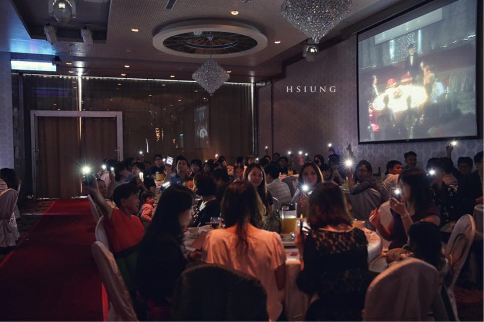 這張婚宴的照片是在一間裝潢華麗的宴會廳,許多賓客圍坐在餐桌旁,舉起手機拍照,場面熱鬧,充滿喜慶的氛圍。