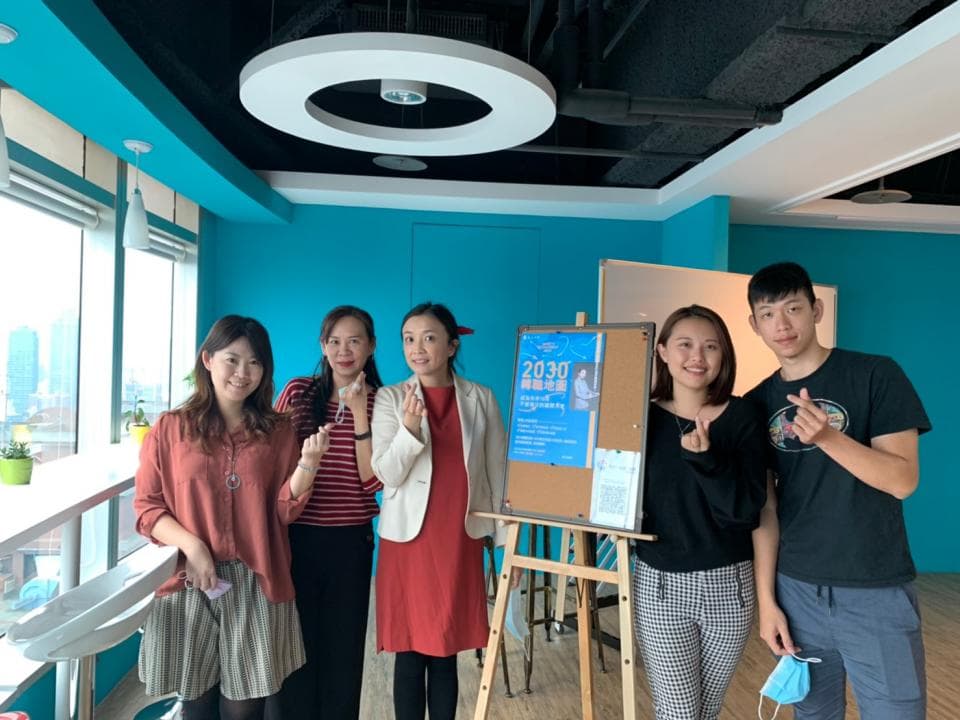 這張照片是五個年輕人在一個明亮的辦公室裡,其中一個穿著紅色洋裝的女生站在一個寫著“2030 願景”的看板前面。