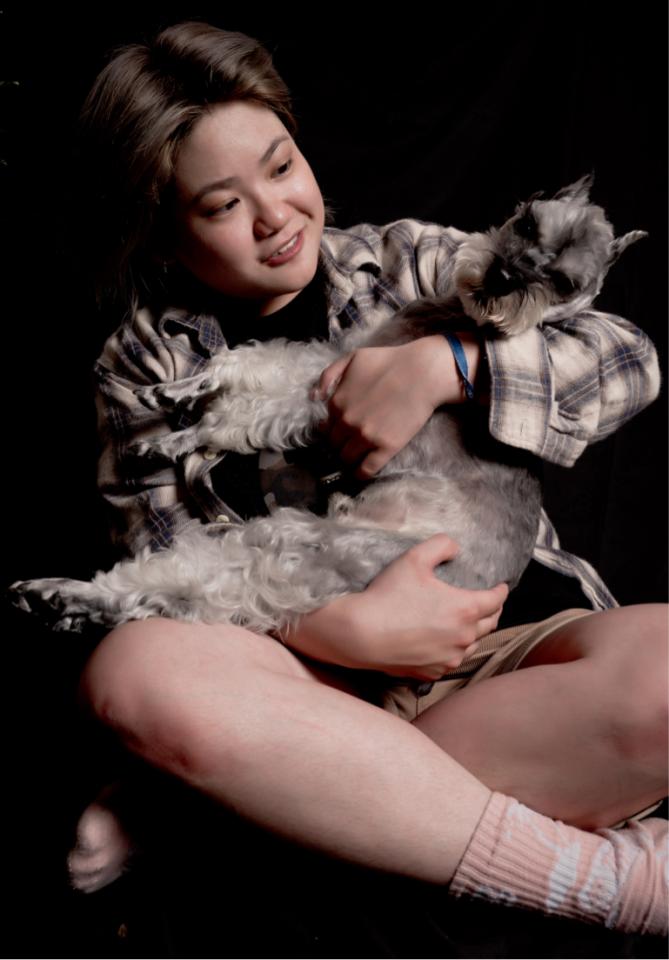 一位年輕女性坐在黑暗背景中,雙手抱著一隻小型犬,她穿著格紋襯衫,狗狗則被抱在她的懷裡