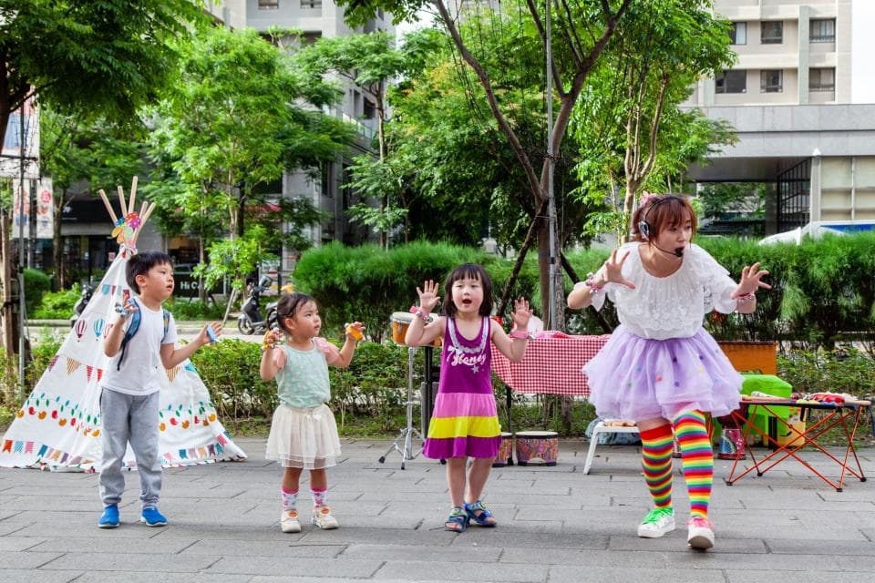 這張照片是一群小朋友在戶外玩耍,其中一個穿著紫色裙子的小女孩正在表演,她非常投入,臉上充滿著笑容,讓周圍的小朋友都看得津津有味。