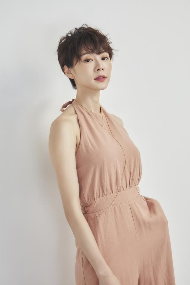 這張照片是一個穿著粉色連身褲的女性。她有著短髮和迷人的笑容,看起來自信又優雅。