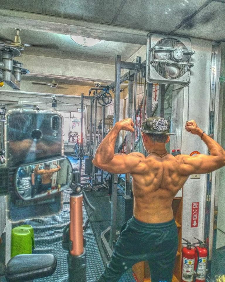 這張「健身房」的照片是一個肌肉發達的男子在健身房做拉背運動,他的背部肌肉線條明顯,穿著運動服裝,背景是健身房的器材。