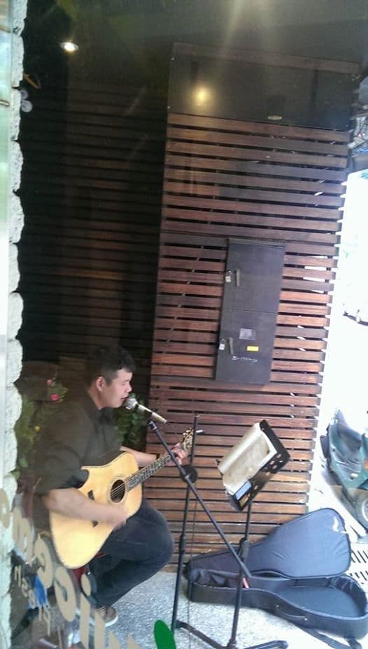 這張照片是一個年輕人正在演奏木吉他的畫面,他穿著簡單的服裝,表情專注,背景是木頭牆壁,他正在演唱,現場非常溫馨,他很享受音樂。