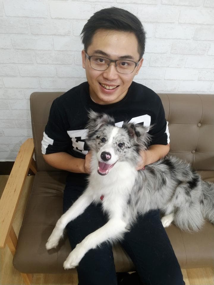 這張照片是一個男人和他的狗在沙發上。男人穿著黑色T恤,戴著眼鏡,狗狗是一隻黑白色的邊境牧羊犬。狗狗把頭放在男人的腿上,男人笑著看著鏡頭。