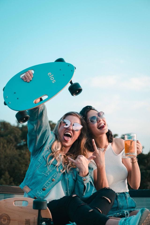 這張「兩個女生在戶外玩滑板」的照片是,一個女生拿著滑板,另一個女生喝著啤酒。