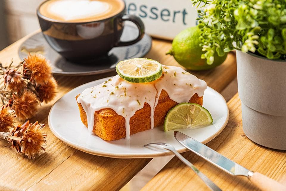 這張「檸檬蛋糕、咖啡和綠色植物」的照片是,一張木質桌子上,放著一杯咖啡、一塊淋上檸檬醬的蛋糕、一盤檸檬和一小盆綠色植物。