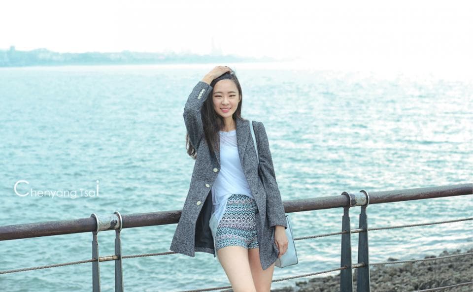 這張照片是一個年輕女子站在海邊,穿著一件灰色的外套和一條印花短褲,她用手撫摸著頭髮,面帶微笑,看著鏡頭。她身後是一片藍色的海洋,天空是晴朗的。
