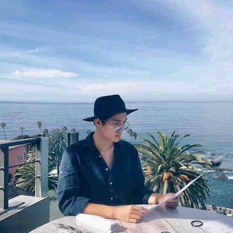 一個戴著黑帽和墨鏡的年輕男子坐在海邊的陽台上,手上拿著一張紙,面向大海,背景是藍色的天空和大海,陽光燦爛