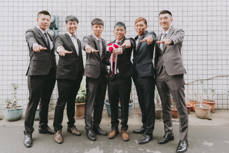 這張照片是六個穿著西裝的男生站在一起,他們都面向鏡頭,其中一位男生手持花束,他們的手指都指向鏡頭,畫面顯得輕鬆愉悅。