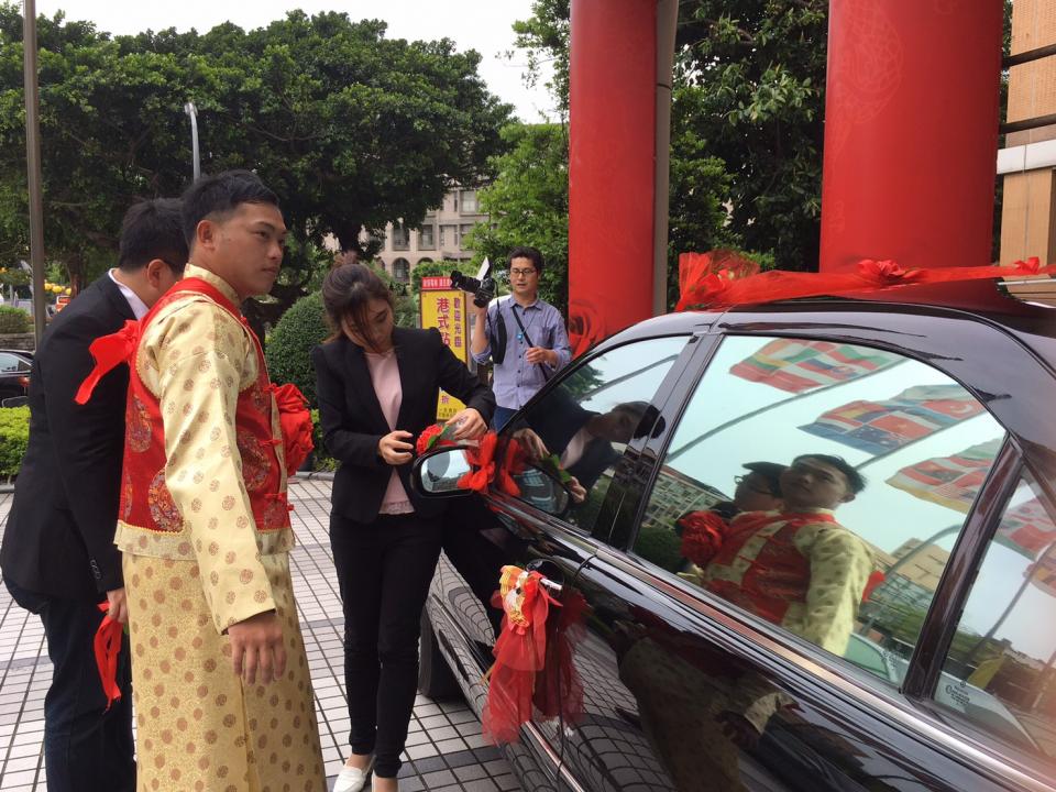 這張「婚禮」的照片是一對新人在汽車前合影,新郎穿著金色傳統服裝,新娘穿著黑色西裝外套和白色襯衫,旁邊還有他們的親朋好友。