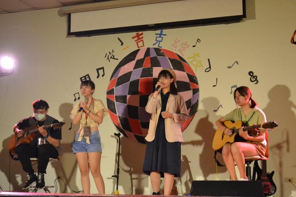 這張照片是四個年輕人在舞臺上表演,他們正在唱歌和彈吉他。其中一位女生穿著粉色外套,手拿麥克風,正在唱歌。她的身旁站著一位女生,穿著黃色短袖和牛仔短褲,手拿麥克風,正在唱歌。舞臺上還有一位男生和女生,他們分別拿著吉他,正在彈奏音樂。這張照片充滿了青春活力,讓人感受到音樂的魅力。