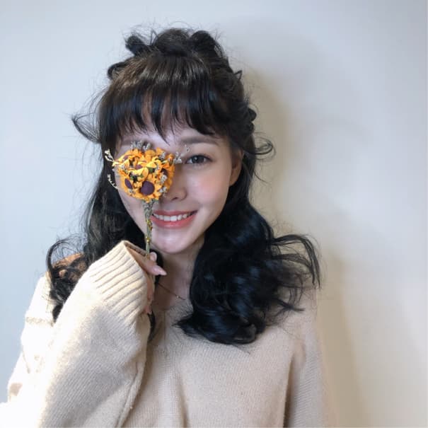 這張照片是一個年輕女性,她拿著一朵枯萎的向日葵遮住了一隻眼睛,露出甜美的笑容。