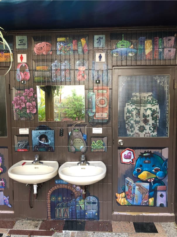 這張「公共廁所」的照片是一個佈滿彩繪的洗手檯,牆壁上裝飾著許多不同的圖案,像是卡通人物、風景畫等,兩邊各有洗手檯和鏡子,看起來很活潑有趣。