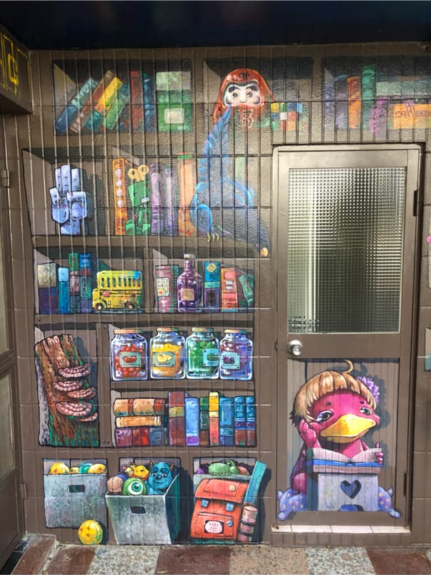 這張「牆壁」的照片是 一個塗鴉作品,上面畫著書架,書架裡放著很多物品,還有卡通人物。