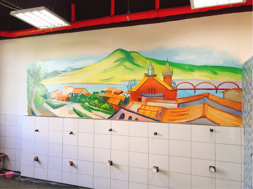 這張「廁所牆壁上畫有鄉村風景」的照片是,一幅色彩繽紛的壁畫,上面描繪著一座小鎮和一座橋,背景是綠色的山丘和水。