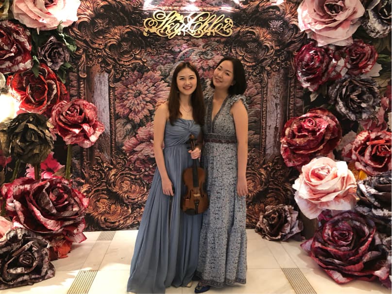 這張「兩個穿著禮服的女生站在裝飾有大量人造花牆壁前」的照片是,其中一個女生拿著小提琴。