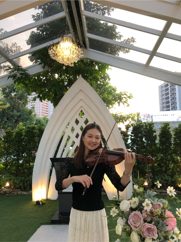 這張照片是一個穿著黑色上衣和白色裙子的年輕女子,在一個戶外婚禮場地演奏小提琴。她站在一個白色拱門前,背景是一片茂盛的綠色植物。