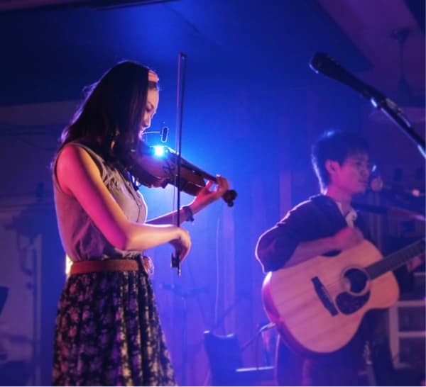 這張照片是兩個年輕人在舞臺上表演。女歌手穿著花裙子,正在演奏小提琴,而男歌手則坐在她旁邊,彈著吉他。他們似乎正在演奏一首歌,享受音樂帶來的樂趣。