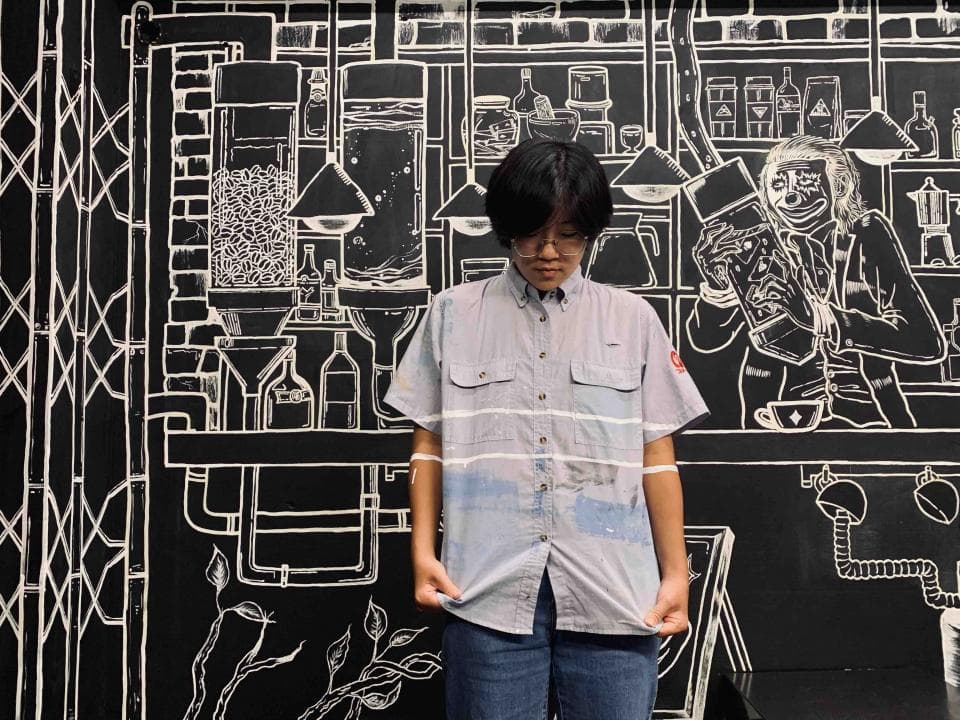 這張照片是一個年輕女子站在一個畫滿圖案的牆壁前面。她穿著一件藍色條紋襯衫,正看著鏡頭。牆上有一幅咖啡館的素描,畫得很逼真。