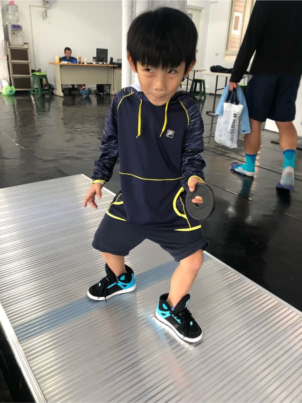 這張照片是一個年輕男孩站在一個金屬板上的照片。他穿著藍色和黃色的運動衫,黑色的褲子和藍色的運動鞋。他右手拿著一個黑色的輪子。