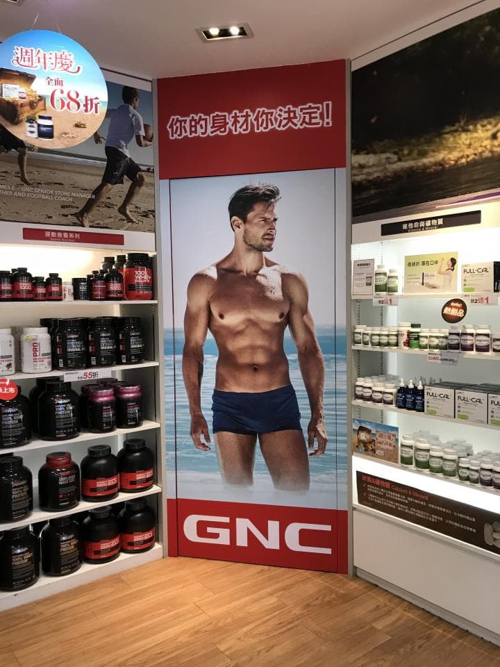 1) 照片顯示一家 GNC 商店的內部,其中一張大型廣告海報展示了一名男性健美運動員,該廣告海報的標題是「你的身材你決定!」