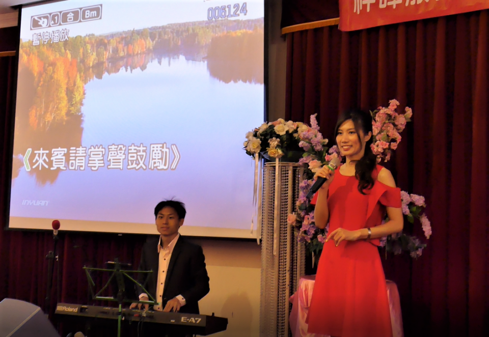 這張照片是一場演唱會,一名身穿紅色洋裝的女性正在舞臺上唱歌,旁邊的男性則在伴奏。