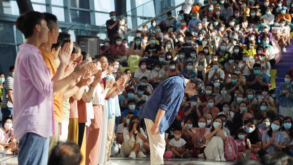 這張「一群人」的照片是一群人在舞臺上表演,臺下觀眾熱情鼓掌。舞臺上有數位表演者,他們正在表演,其中一位表演者向觀眾深深鞠躬,以感謝觀眾的掌聲。舞臺下有很多觀眾,他們都戴著口罩,並熱情地鼓掌。