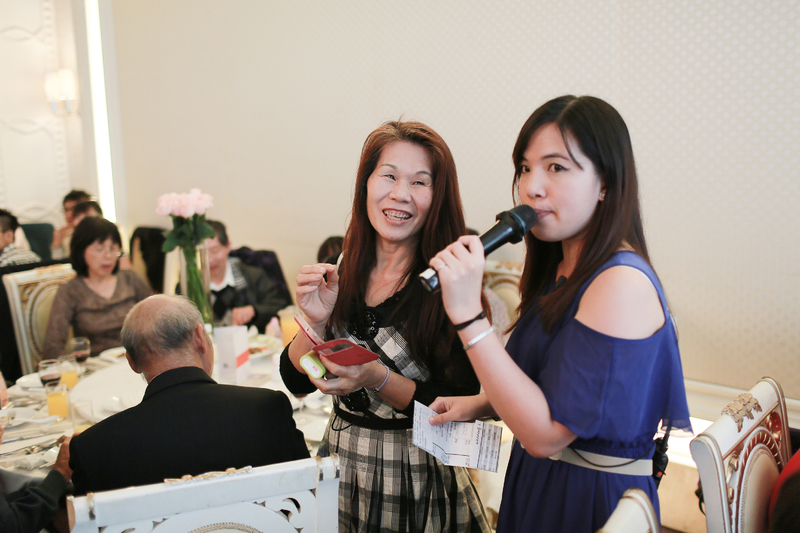 這張「婚禮」的照片是兩位女性在婚禮上說話,其中一人手持麥克風,另一位則手持紅色的卡片。她們的背景是一群賓客,還有白色桌布和花卉裝飾。