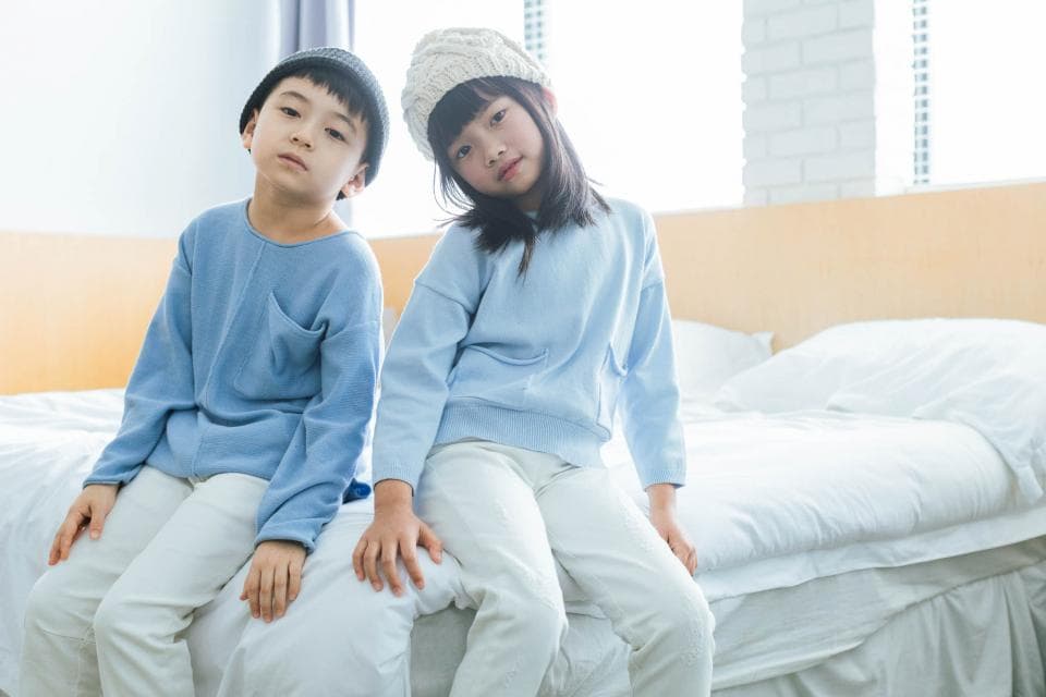 這張「兩個孩子坐在床上的」照片是,一個戴著灰色帽子的男孩和一個戴著白色帽子的女孩,他們都穿著藍色的衣服,坐在一張白色床單的床上。