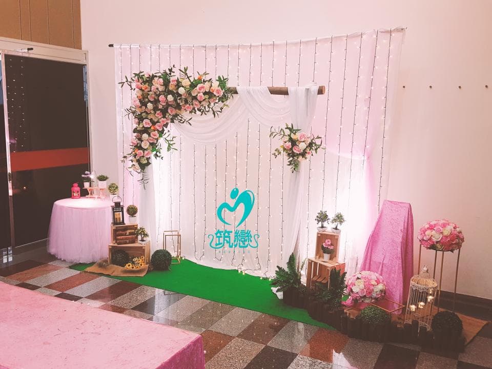 這張「婚禮佈置」的照片是一個婚禮現場,背景是一個白色布幔,上面掛滿了燈泡,兩旁擺放了花束,中間有一個木製的拱門,拱門上也掛上了白色布幔,地上鋪著綠色的地毯。