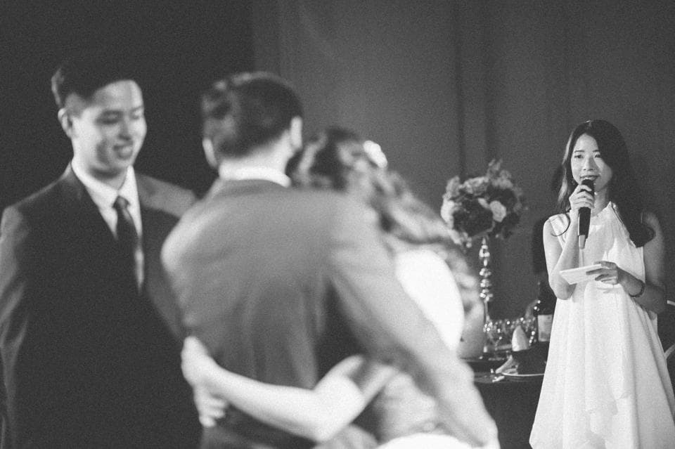 這張婚禮的照片是一對新人,新郎穿著西裝,新娘穿著白色禮服,旁邊站著一個女人正在臺上講話。