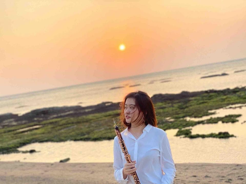 這張照片是一個年輕女子在沙灘上吹奏雙簧管。她穿著白色襯衫,頭髮披散著,背景是夕陽西下的海灘。
