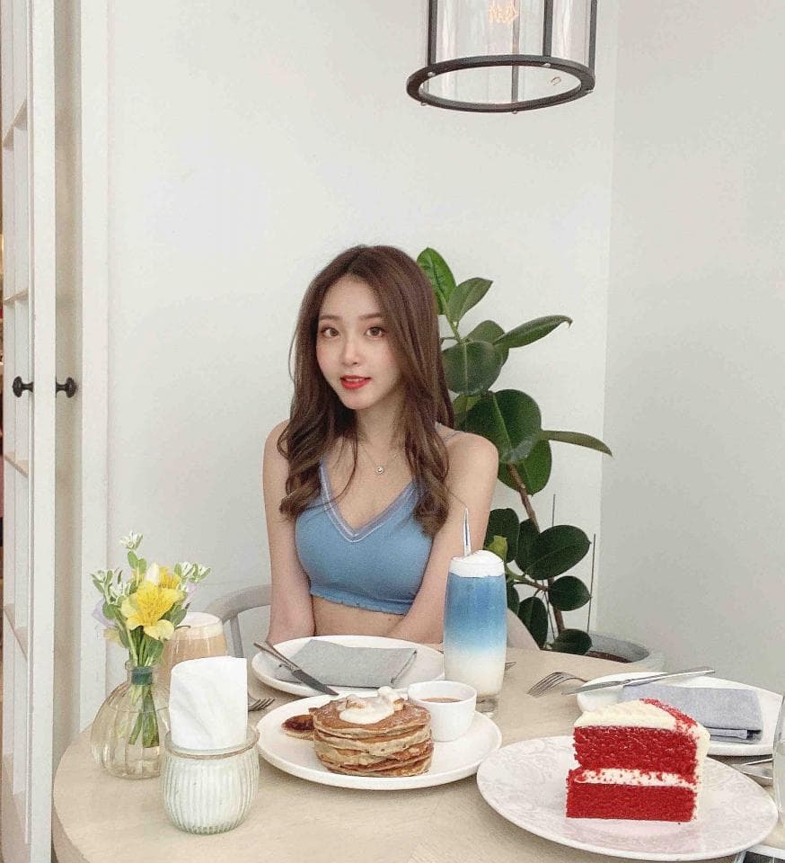 這張照片是一名年輕女子在餐桌上吃飯的樣子,她穿着藍色的吊帶上衣,背景是白色牆壁和綠色植物,桌上放着蛋糕和飲品。