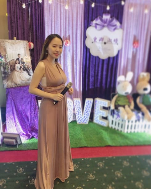 這張照片是一個穿著淺棕色禮服的女性,她拿著麥克風,站在一個婚禮佈置的背景前,背景上有紫色亮片布、LOVE字樣燈飾和可愛的兔子玩偶。