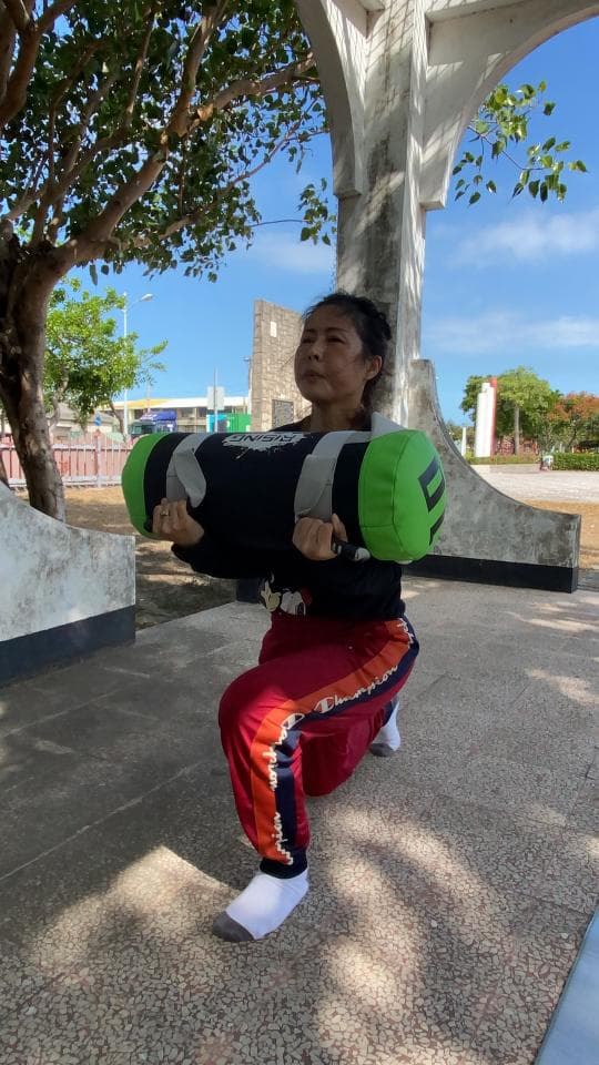 這張照片是一個年輕女子正在舉重。她舉著一個綠色和黑色的沙袋,雙手緊握,做著深蹲動作。