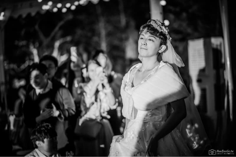 這張照片是一位身穿婚紗的男性站在人群中,他頭戴頭紗,神情愉悅,顯得十分喜悅。他周圍的環境昏暗,但他的婚紗在燈光下顯得格外耀眼。