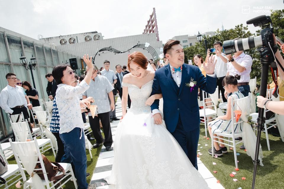 這張照片是一對新人正在舉行婚禮,新娘穿著白色婚紗,新郎穿著藍色西裝,他們正走向觀禮的賓客,背景是一片綠色的草坪。