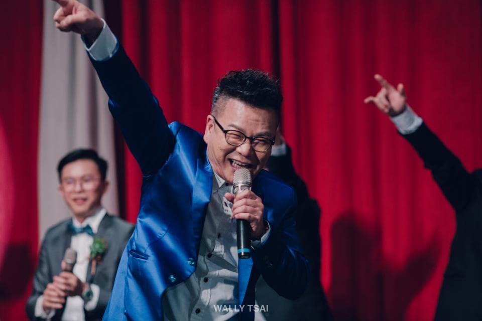 這張照片是一個男人正在臺上拿著麥克風唱歌,他穿著藍色西裝,神情興奮,背景是一片紅色布幕。