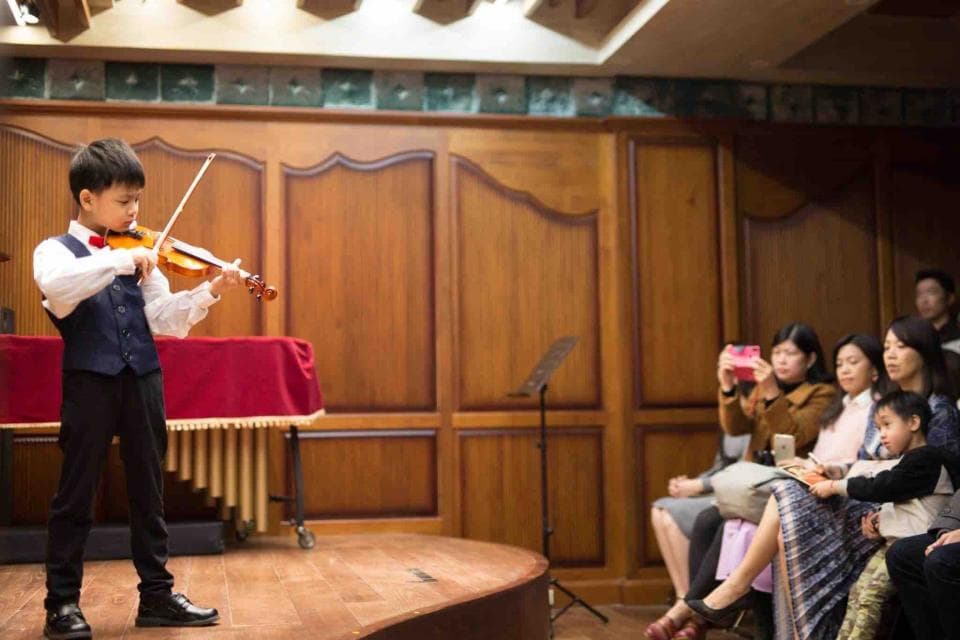 這張照片是一個年輕的小提琴手正在舞臺上表演,他的面前放著一把小提琴和一個小巧的木琴。他專注地拉著小提琴,看起來非常認真,而臺下有許多觀眾正在欣賞他的演出。