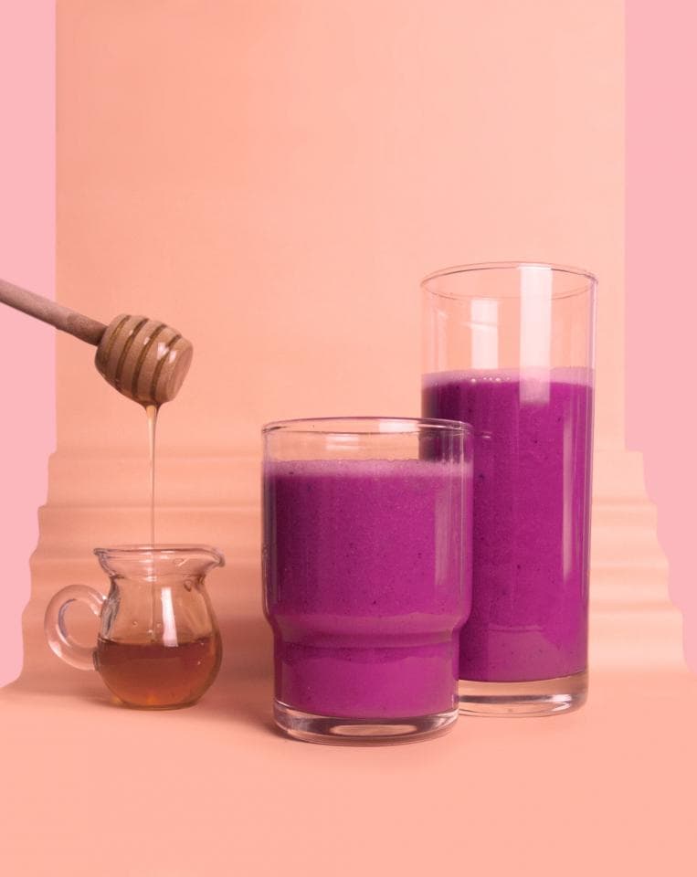 這張「兩杯紫色飲品和蜂蜜」的照片是,兩杯紫色飲品放在粉色背景上,其中一杯較高,另一杯較矮。蜂蜜從木製蜂蜜棒滴下,旁邊有一小罐蜂蜜。