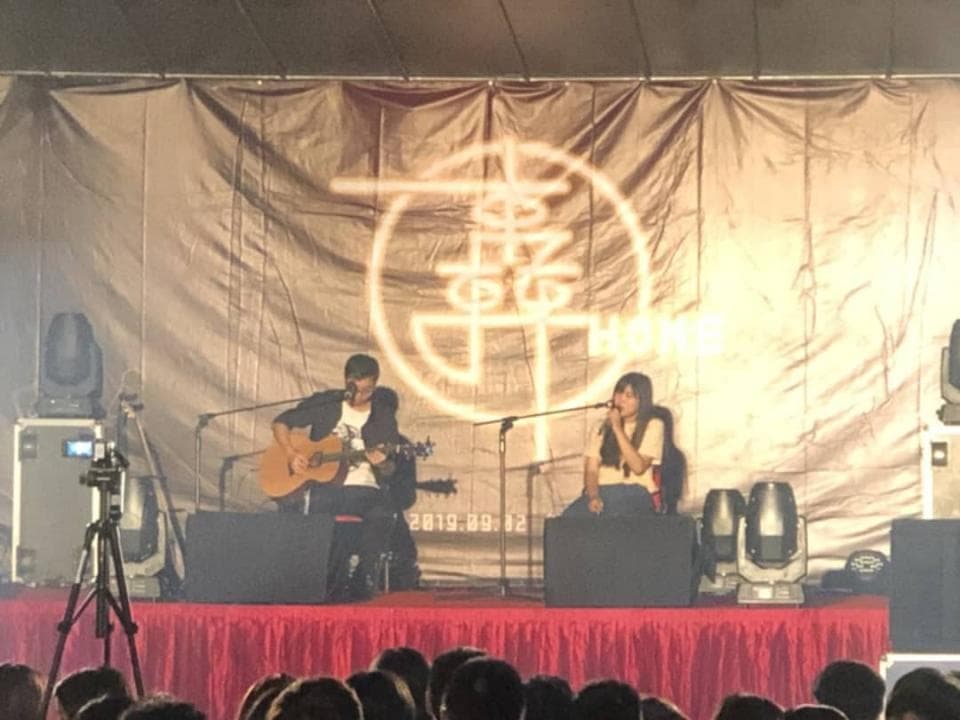 這張照片是兩個歌手正在台上表演,背景的布幕上有個大大的金色圓圈,裡面寫著「幸福」。