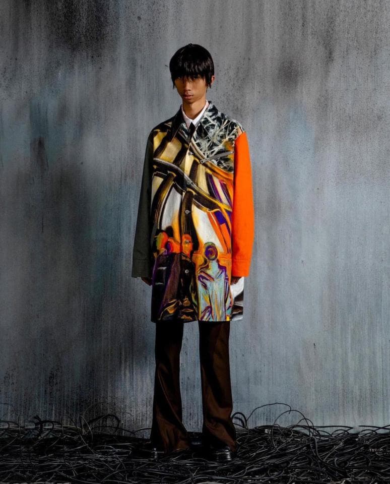 這張照片是一個穿著印花外套的年輕男子,他站在一個灰色的背景前,背景上有一些黑色的線。