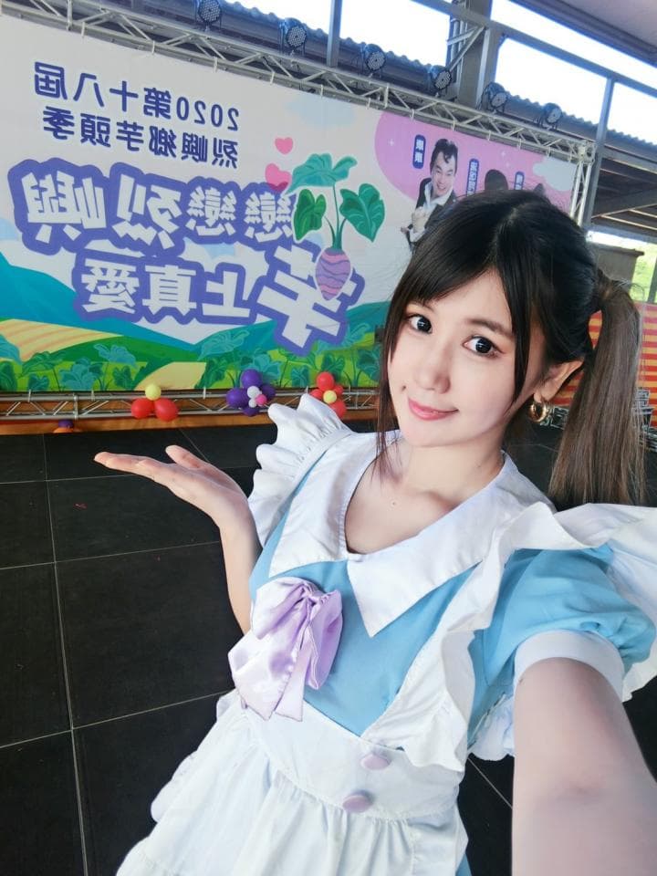 這張「女僕裝」的照片是一位穿著藍色和白色女僕裝的女性。她站在一個背景佈置的場景,看起來像是一個活動或展覽。她微笑著,雙手放在前面,看起來很可愛。